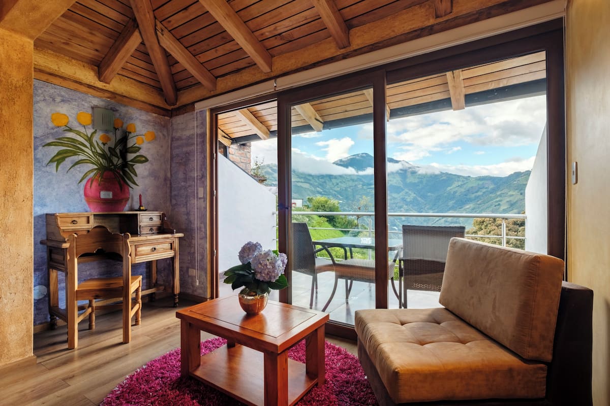 A room with a wooden ceiling, a wooden desk with drawers, a wooden chair, a sofa, a table with a vase of flowers, and a large sliding glass door leading to a balcony with a mountain view.