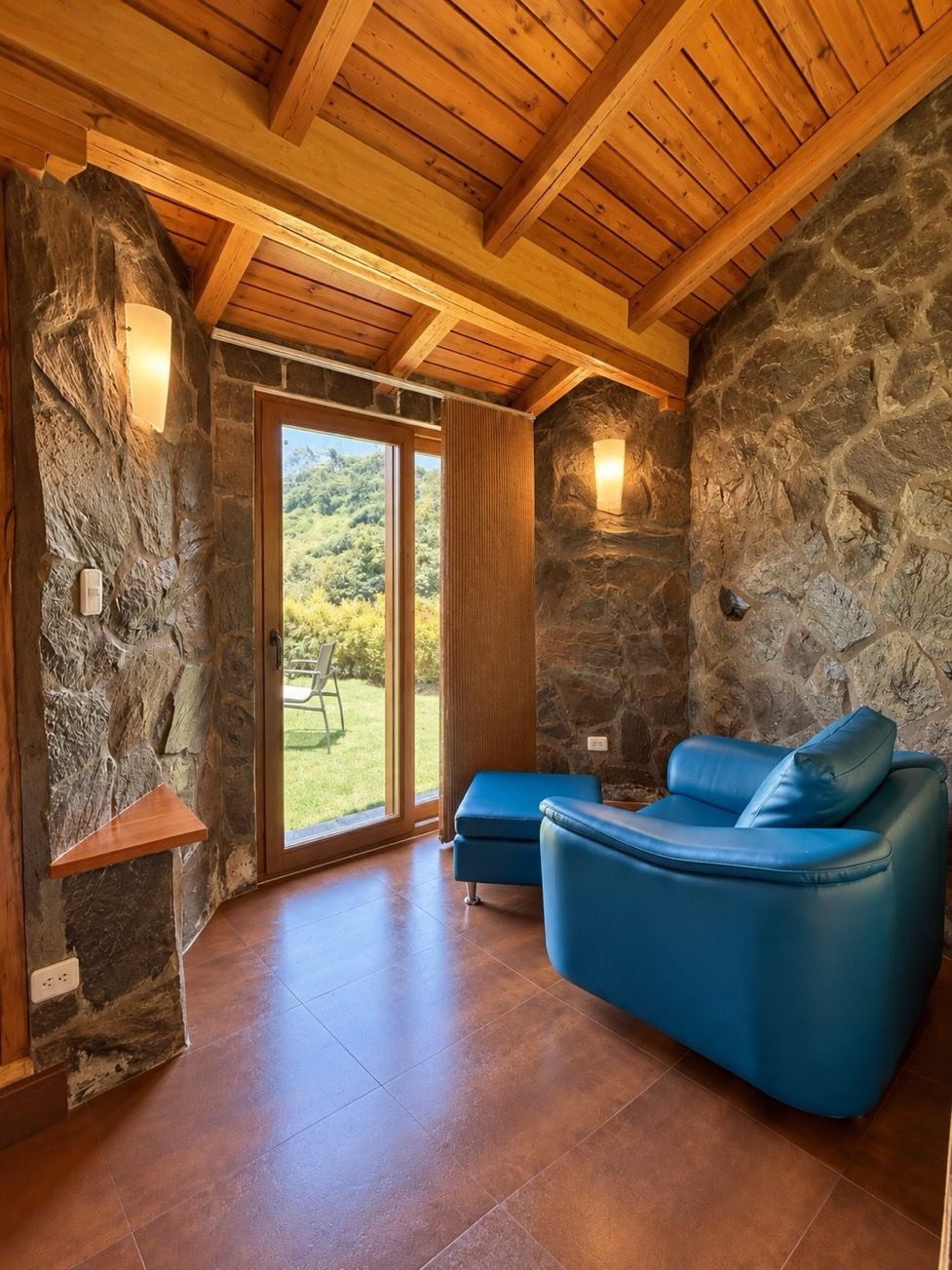 A room with a stone wall, wooden ceiling beams, a blue leather sofa, and a view of greenery outside.