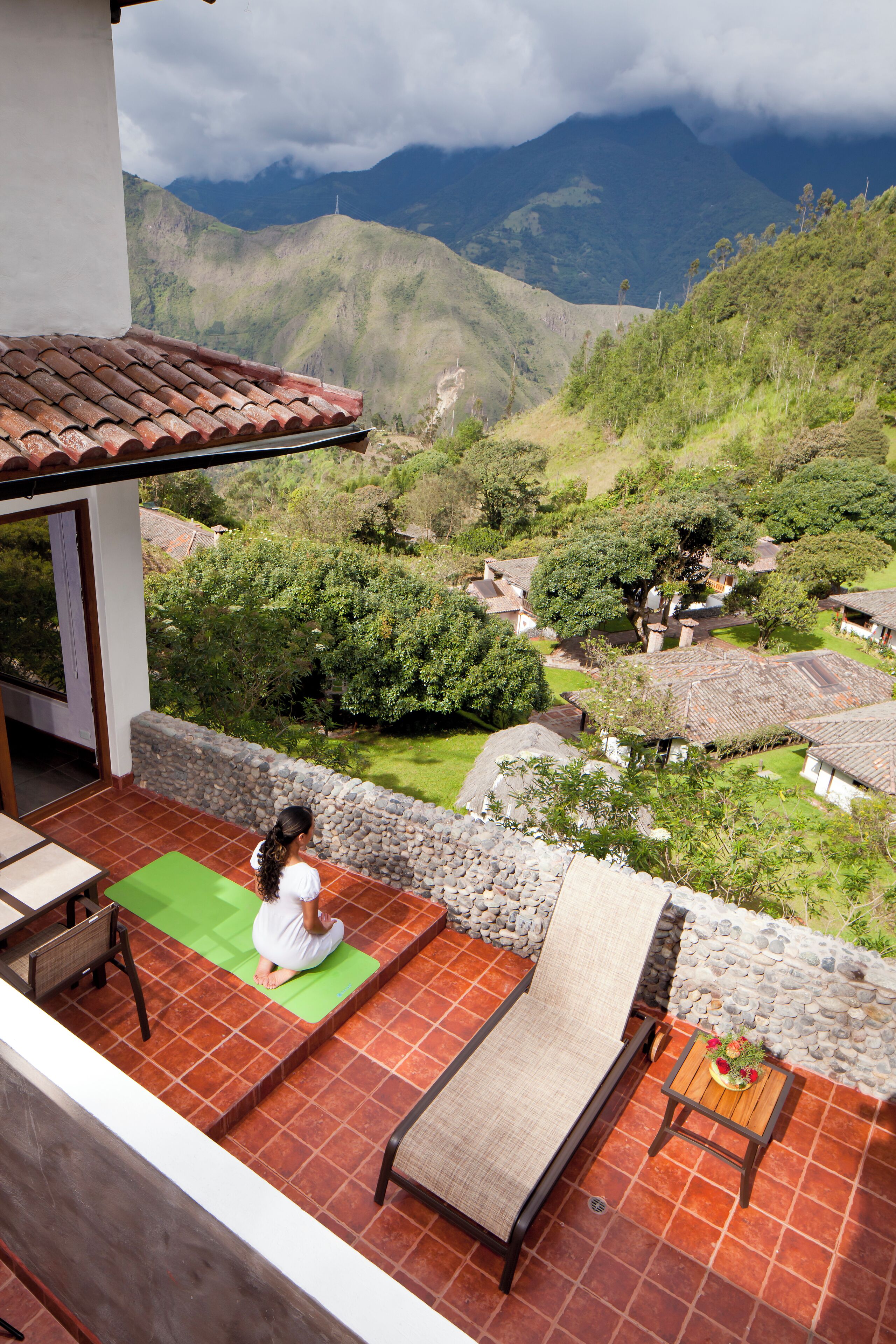 A balcony with a person meditating on a green mat, overlooking a mountainous landscape with traditional houses.
