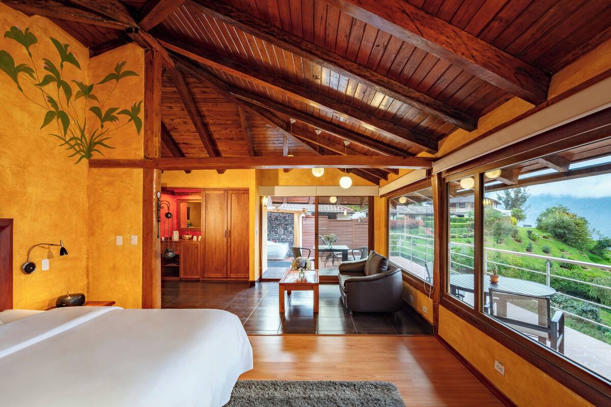 A bedroom with a large bed, wooden ceiling beams, a view of a dining area, and a balcony with a scenic view.