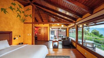 A bedroom with a large bed, wooden ceiling beams, a view of a dining area, and a balcony with a scenic view.