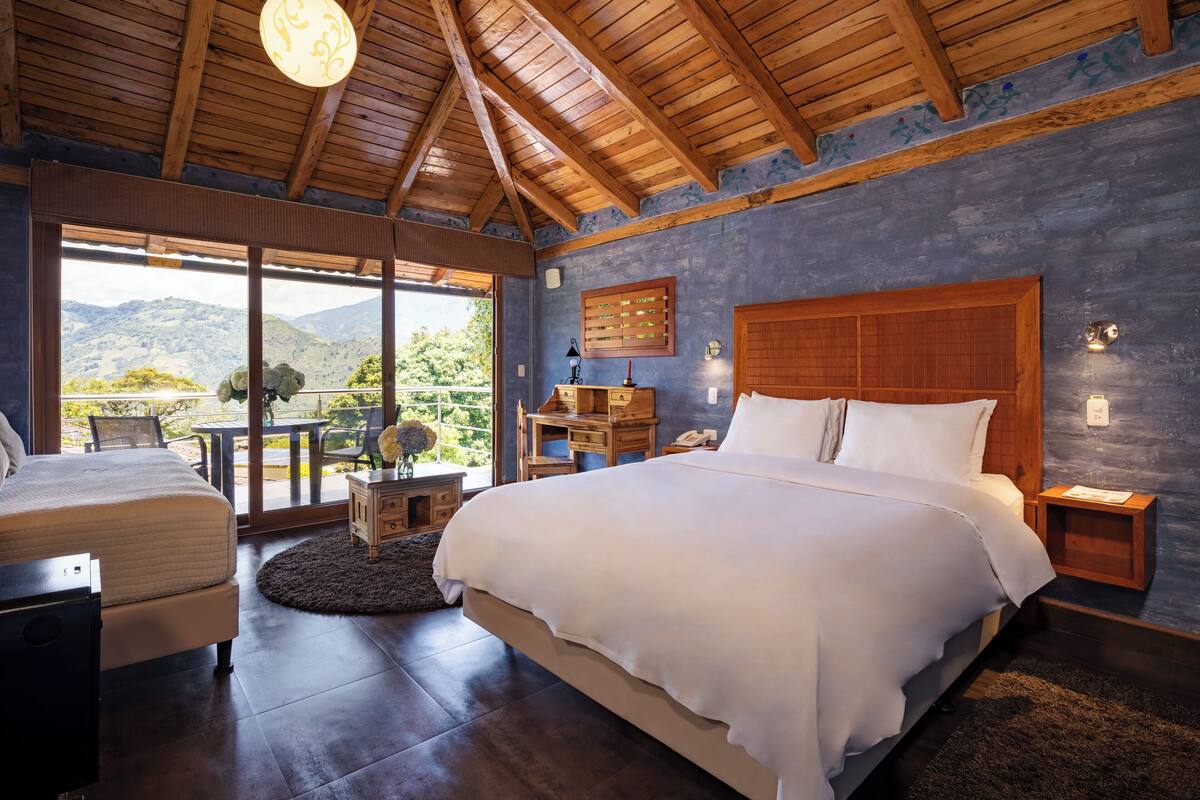 A spacious bedroom with a large bed, wooden ceiling beams, a nightstand, a desk, and a view of the outdoors.
