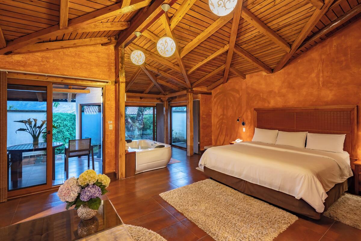 A spacious bedroom with a large bed, wooden ceiling beams, and a view through glass doors to an outdoor area with a hot tub and patio furniture.