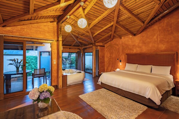 Une chambre spacieuse avec un grand lit, des poutres apparentes en bois au plafond et une vue sur l’extérieur, accessible par des portes vitrées, donnant sur un espace extérieur aménagé avec un jacuzzi et du mobilier de patio.