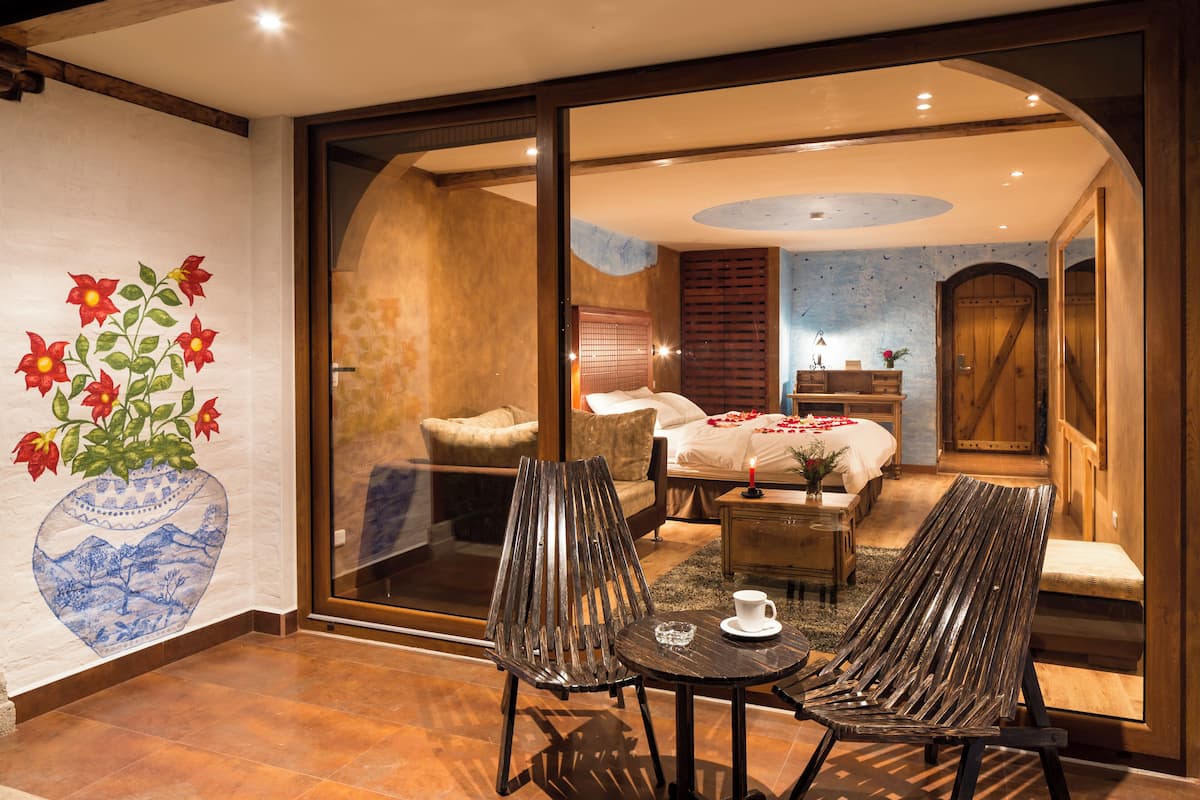 A room with a bed, a wooden bench, a small table, and a vase with flowers on the wall.
