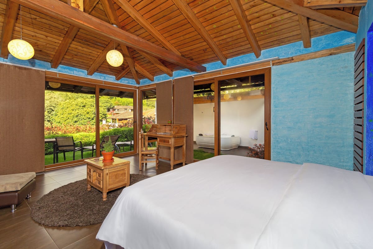 A bedroom with a large bed, wooden furniture, and a view of a garden through glass doors.