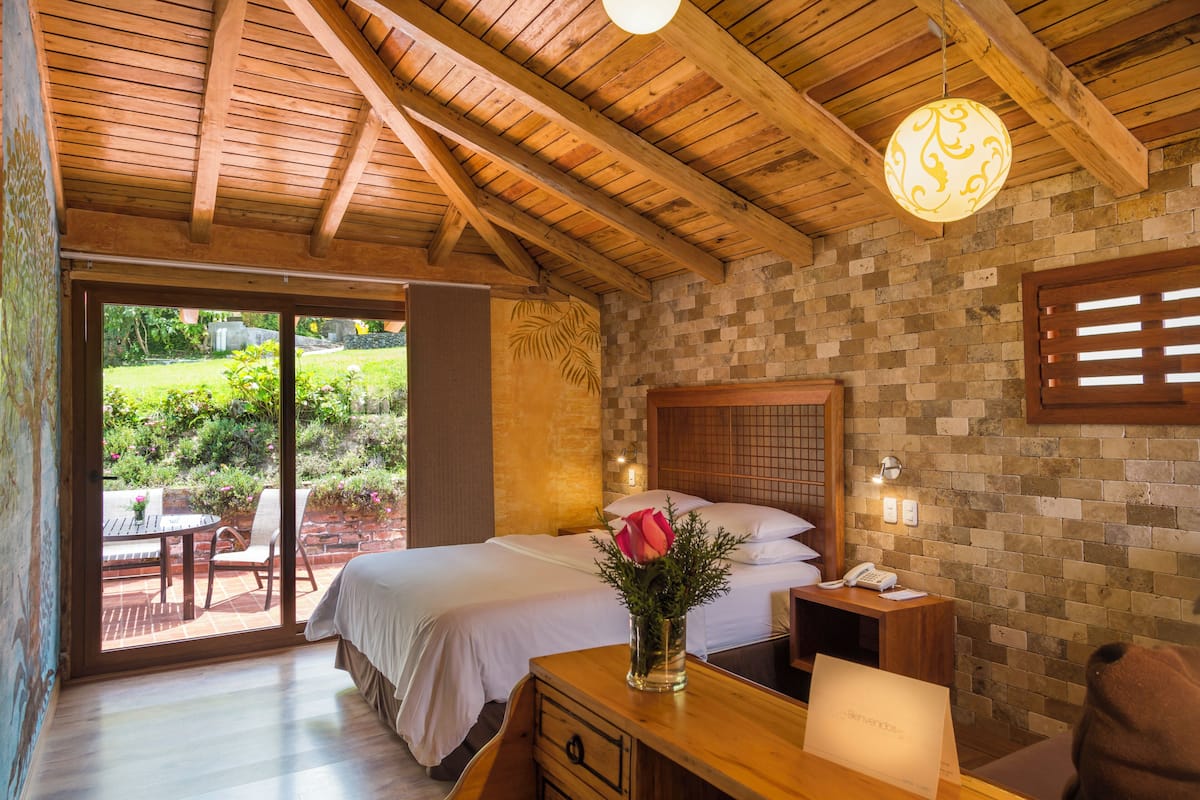 A bedroom with a wooden ceiling, a brick wall, a bed with white linens, a bedside table with a vase of flowers, and a view of a garden through a sliding door.