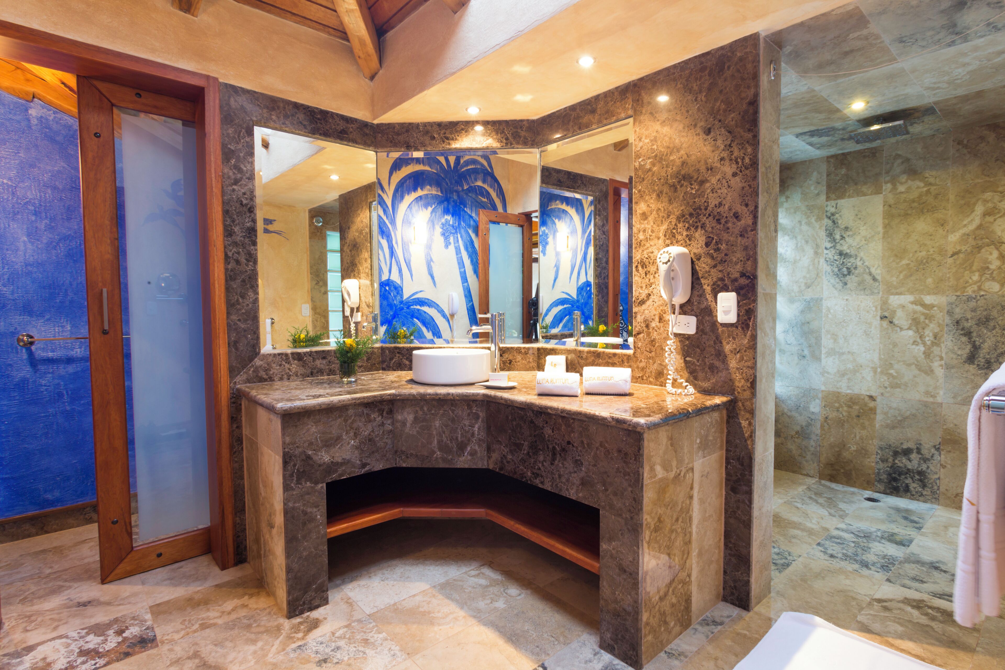 A bathroom with a marble vanity, a large mirror with a palm tree design, a shower area, and a blue door.