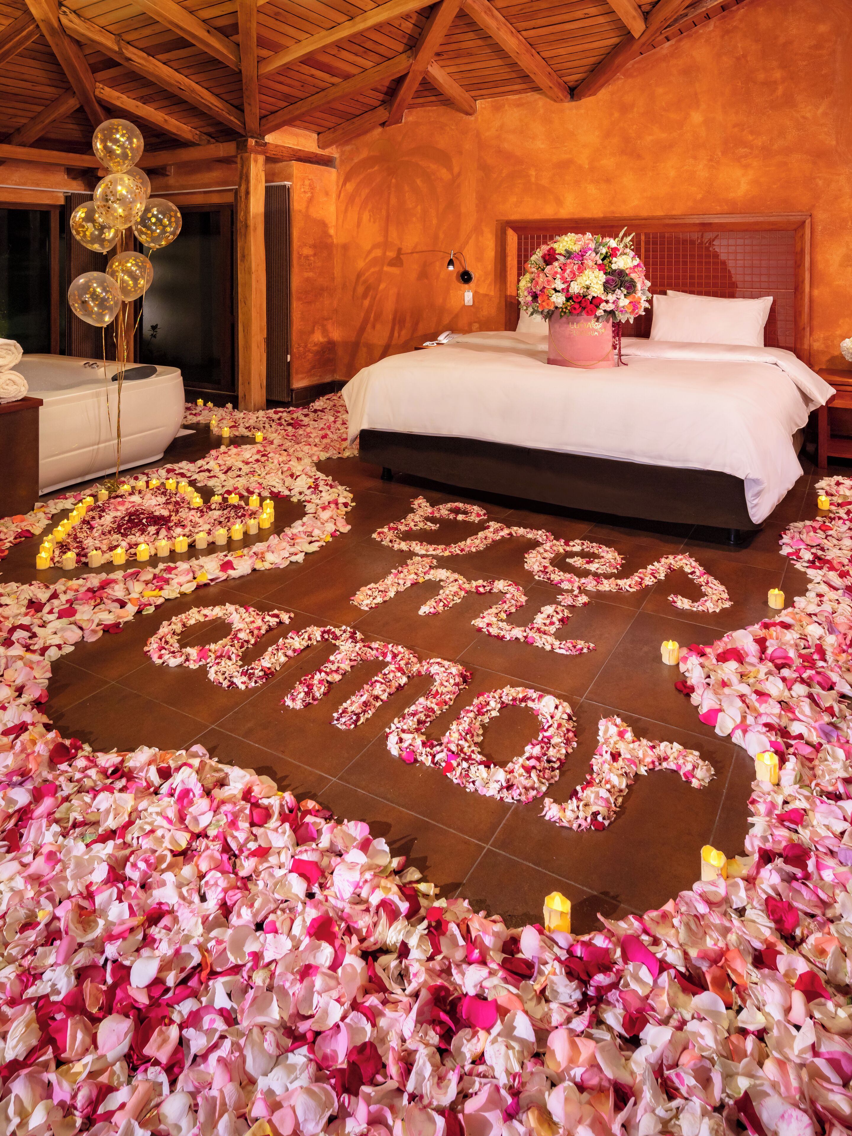A hotel room with a bed, a bedside table, a nightstand, a decorative flower arrangement, and a wall with a wooden ceiling.