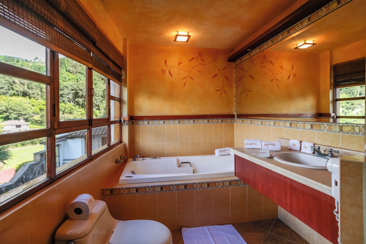 Un baño con bañera, inodoro y lavamanos. Hay ventanas y se ve vegetación afuera.