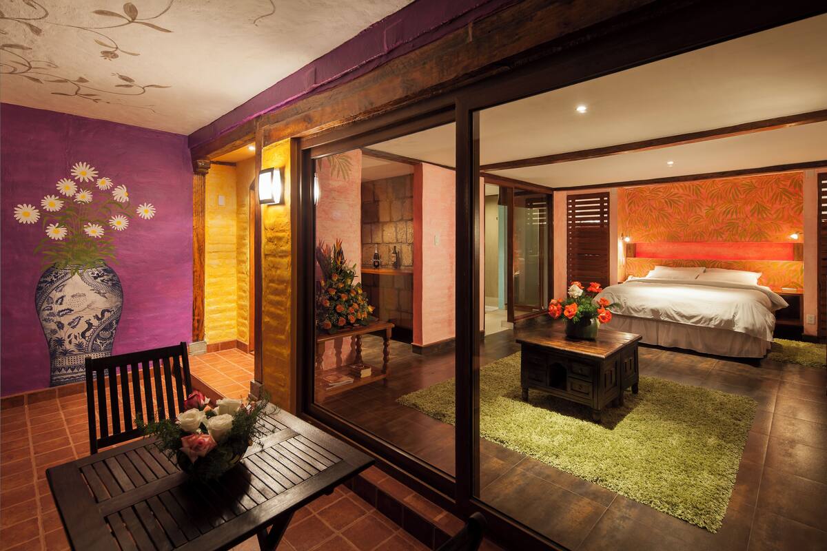A modern hotel room with a large bed, a small table with flowers, a bench, and a view through a glass door to another room.