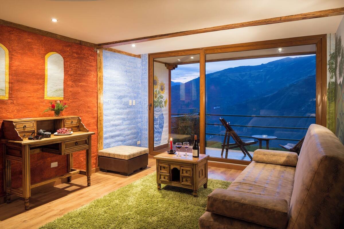 A cozy room with a wooden desk, a cushioned bench, and a sofa. A large window offers a view of mountains.