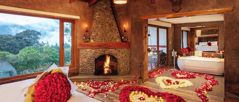 Una habitación con chimenea de piedra, una cama con pétalos de rosa y velas, y vistas al exterior.