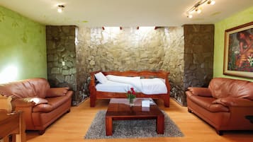 A room with a stone wall, a bed, two leather sofas, a coffee table with a vase of flowers, and a painting on the wall.