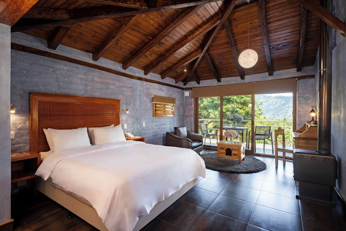 A bedroom with a large bed, wooden ceiling beams, a stone wall, and a view of the outdoors.