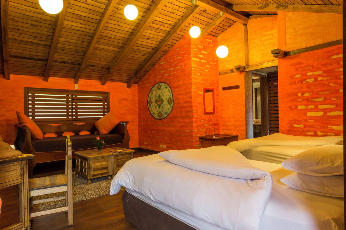 A cozy room with a wooden ceiling, a brick wall, a bed with white bedding, a sofa with orange cushions, and a small table with a vase.