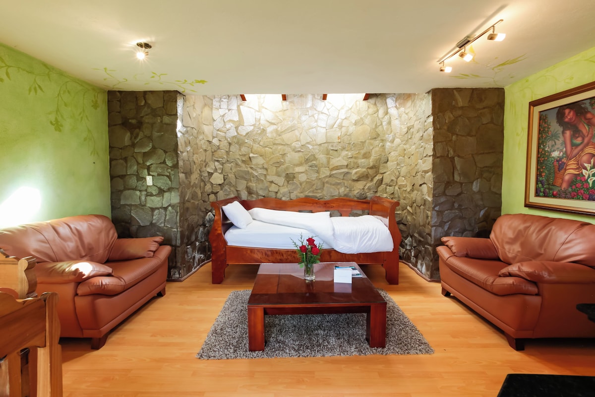 A room with a stone wall, a bed, two leather sofas, a coffee table with a vase of flowers, and a painting on the wall.