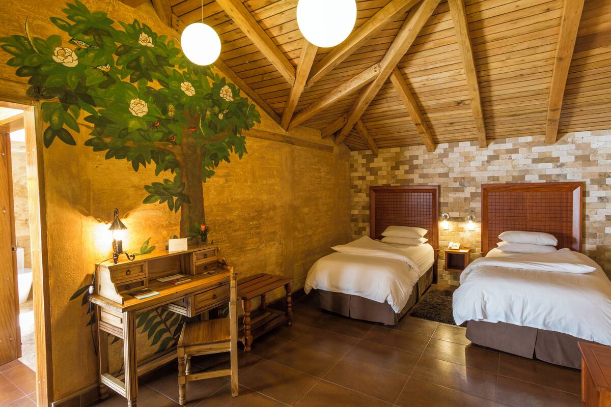 A rustic room with a wooden ceiling, two beds, a desk, and a wall-mounted lamp.