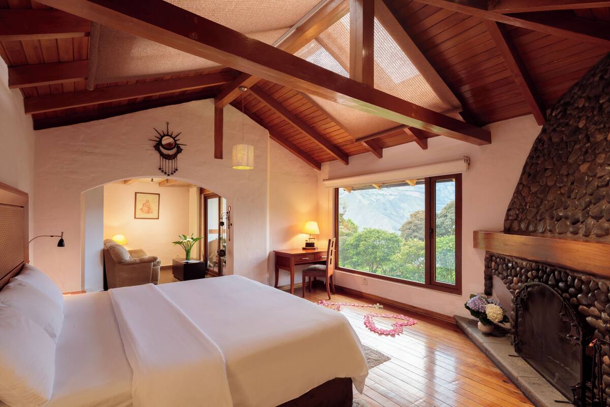 A spacious bedroom with a large bed, a stone fireplace, and a view of the mountains through a large window.