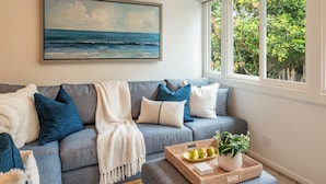 A living room with a grey sofa, blue and white cushions, a painting of a beach, and a window with a view of greenery.