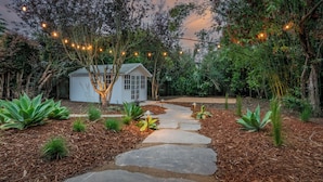 A stone pathway leading to a white shed surrounded by lush greenery and string lights.