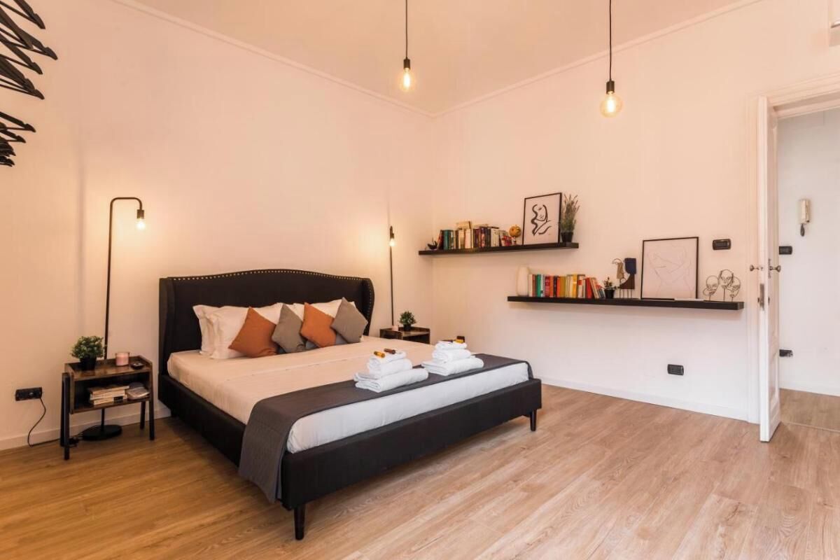 A modern bedroom with a large bed, bedside tables, a bedside lamp, a shelf with books and decorative items, and a door.