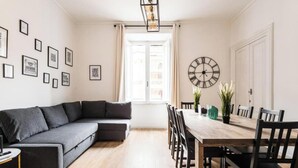 Un salon moderne avec un canapé d’angle gris, une table à manger en bois et des chaises noires, ainsi qu’une grande horloge murale.