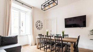 Un coin repas avec une table et des chaises en bois, un téléviseur à écran plat fixé au mur et une grande fenêtre avec des rideaux.
