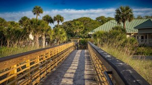 A wooden boardwalk leading to a building with a green roof, surrounded by palm trees and vegetation. - Villa w/ Golf Views! Pool, Near Beach Hilton Head (Hilton Head Island)