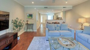 A living room with a sofa, coffee table, and a TV. There is a plant, a lamp, and a kitchen in the background. - Villa w/ Golf Views! Pool, Near Beach Hilton Head (Hilton Head Island)