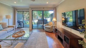 A modern living room with a glass coffee table, a sofa, a chair, a TV, and a sliding glass door leading to an outdoor area. - Villa w/ Golf Views! Pool, Near Beach Hilton Head (Hilton Head Island)