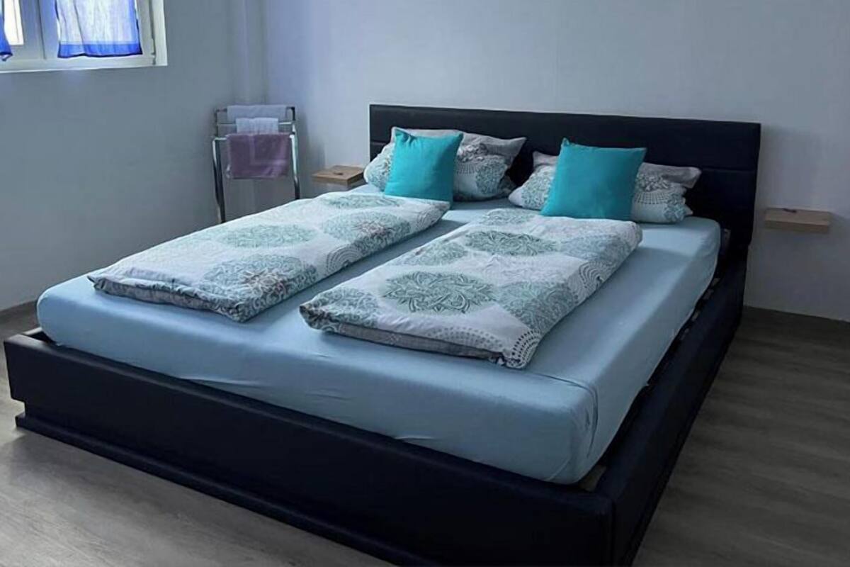 A neatly made bed with a patterned bedspread and pillows, a bedside table with a lamp, and a window with curtains.