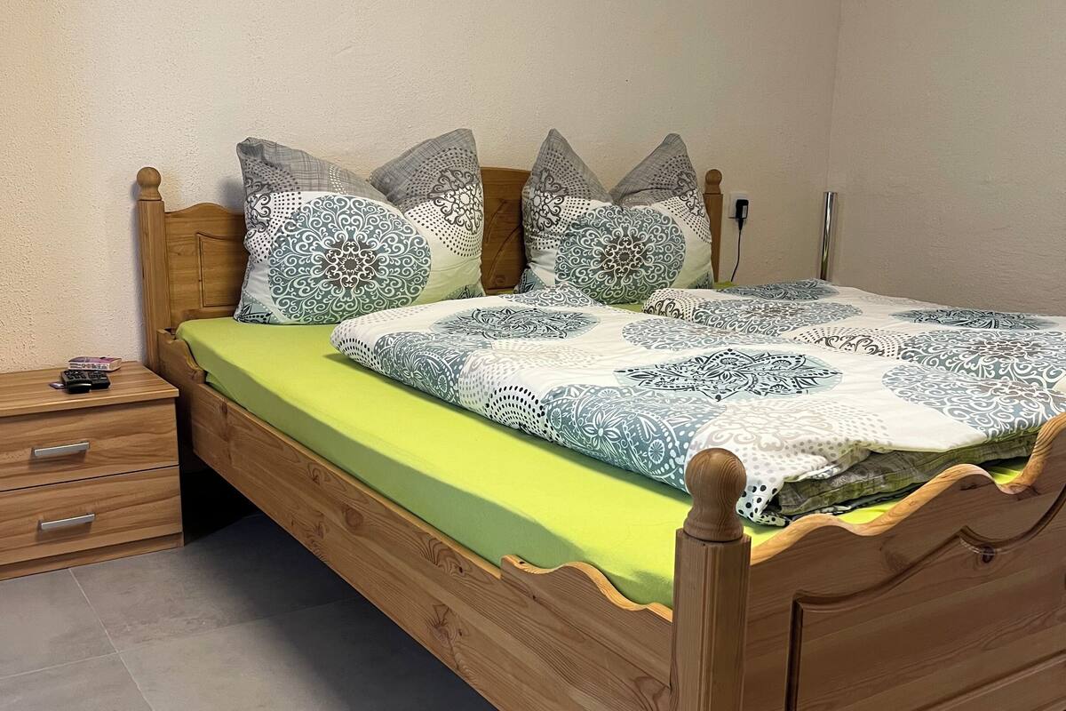 A wooden bed with a green mattress and patterned bedding, a wooden nightstand with a remote control, and a tiled floor.
