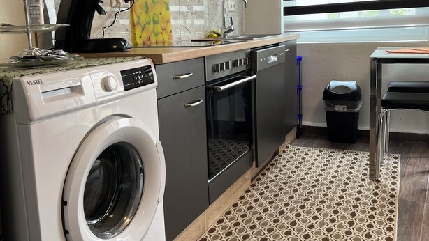 A modern kitchen with a built-in washer and dryer, a coffee machine, and a window with blinds. - Apartments & Zimmer (Vettweiß)