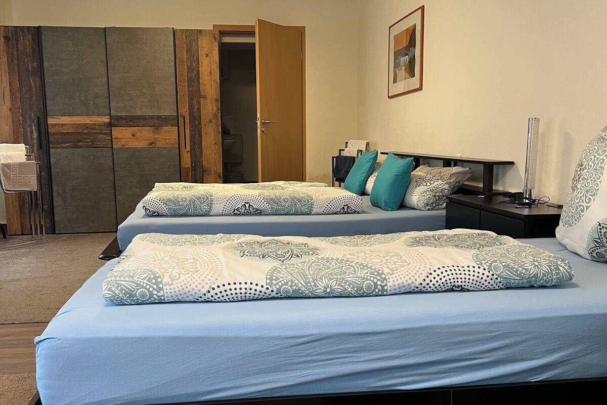 A bedroom with two beds, a green ottoman, a bedside table with a lamp, and a wardrobe.