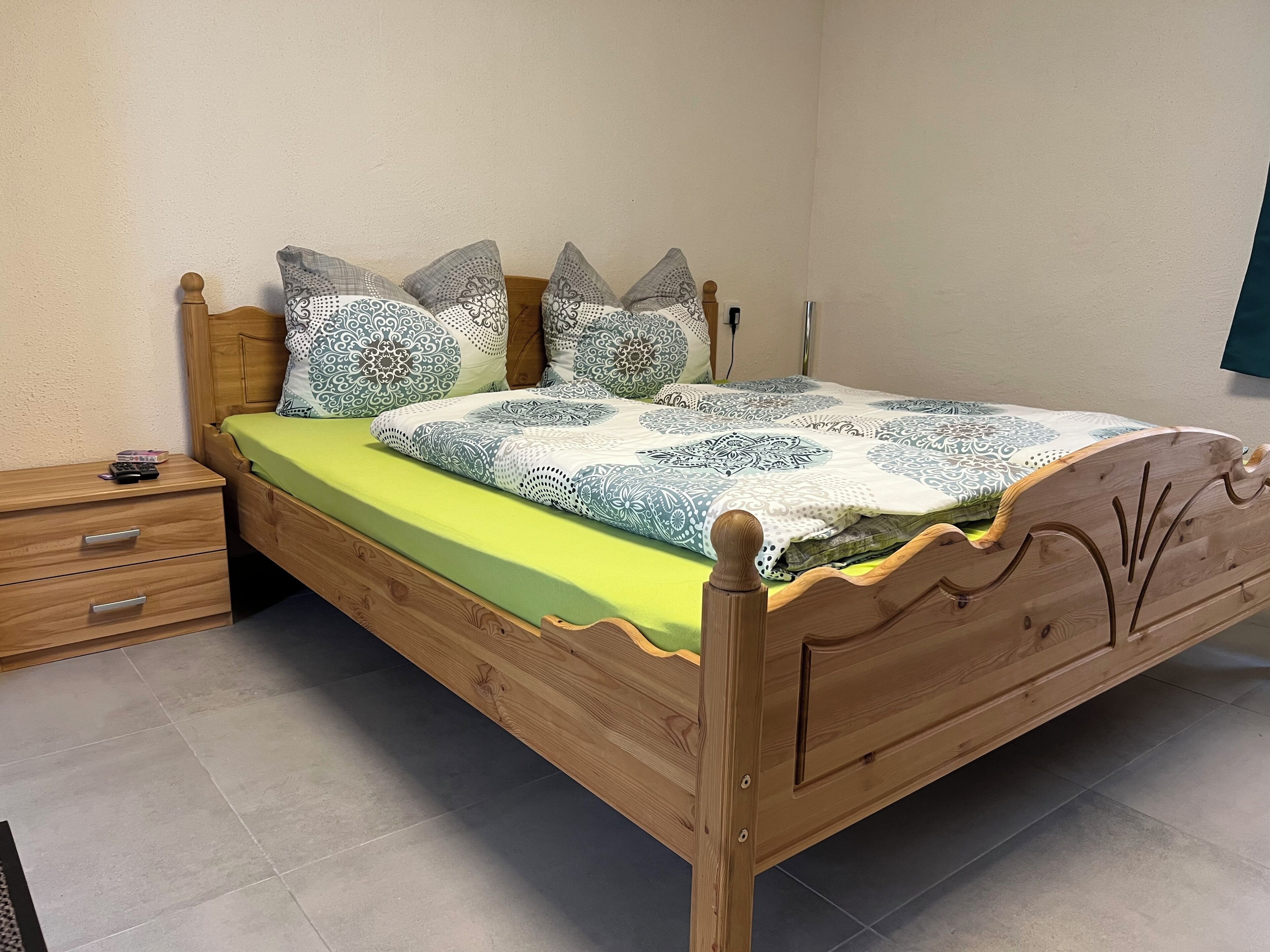 Ein hölzernes Bett mit einer grünen Matratze und gemusterten Bettwäsche, ein Nachttisch aus Holz mit Schubladen und ein Fliesenboden.