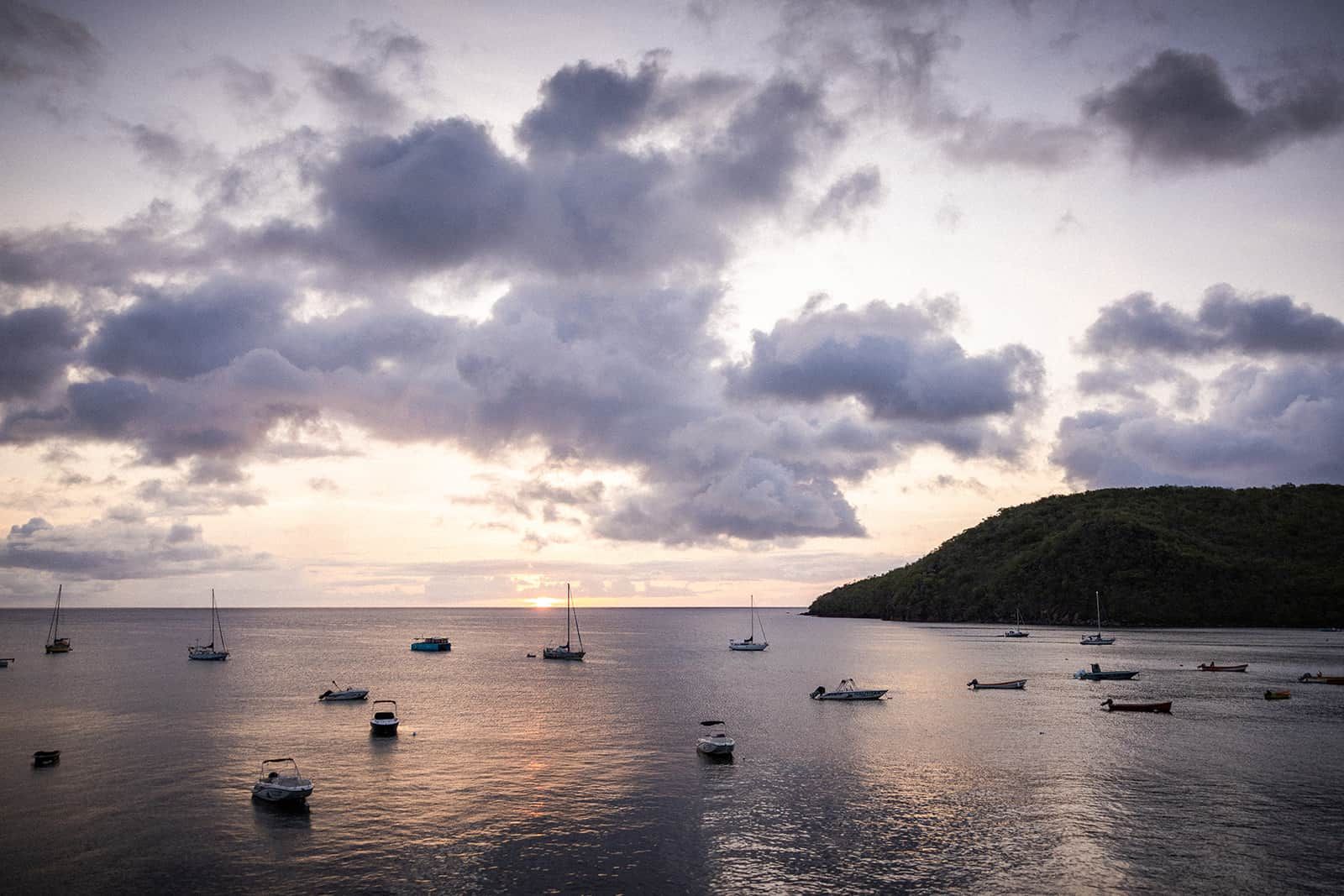 A calm sea with multiple boats anchored, a sunset in the background, and a hilly landscape.