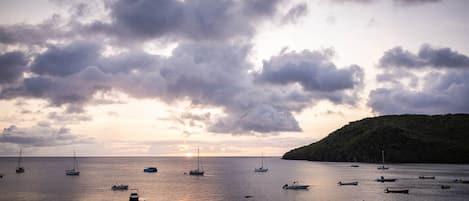 A calm sea with multiple boats anchored, a sunset in the background, and a hilly landscape.