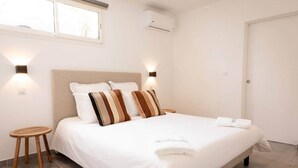 Una cama bien tendida con ropa blanca y almohadas a rayas, un taburete de madera y un equipo de aire acondicionado de pared.