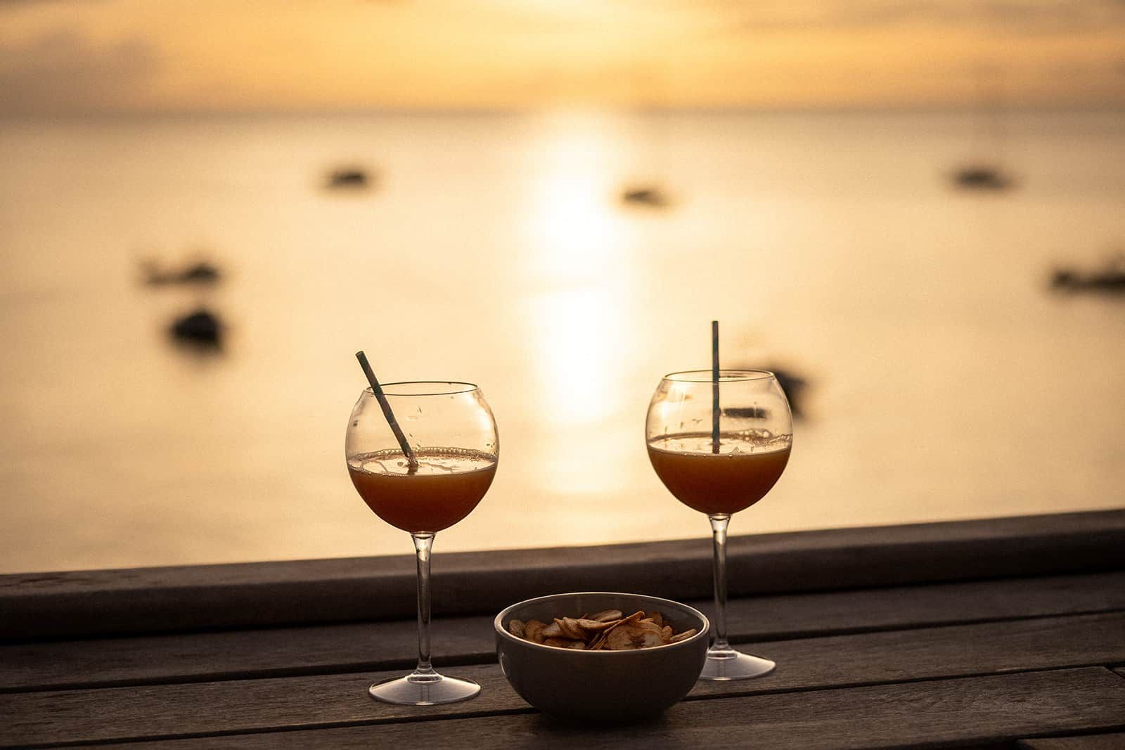 Two glasses of a beverage with straws on a wooden surface, a bowl of snacks, and a view of a body of water with boats.