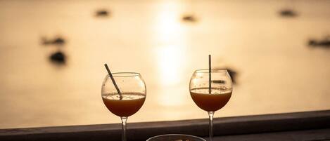 Two glasses of a beverage with straws on a wooden surface, a bowl of snacks, and a view of a body of water with boats.