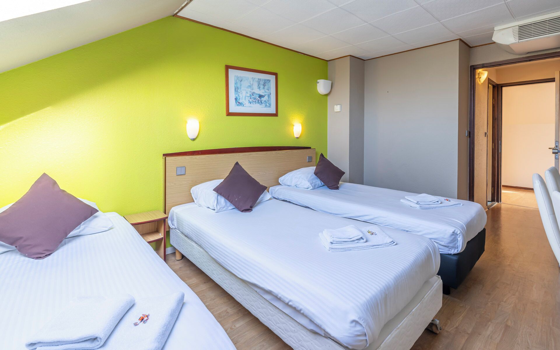 A hotel room with two beds, green walls, a framed artwork, wall-mounted lights, and a door.