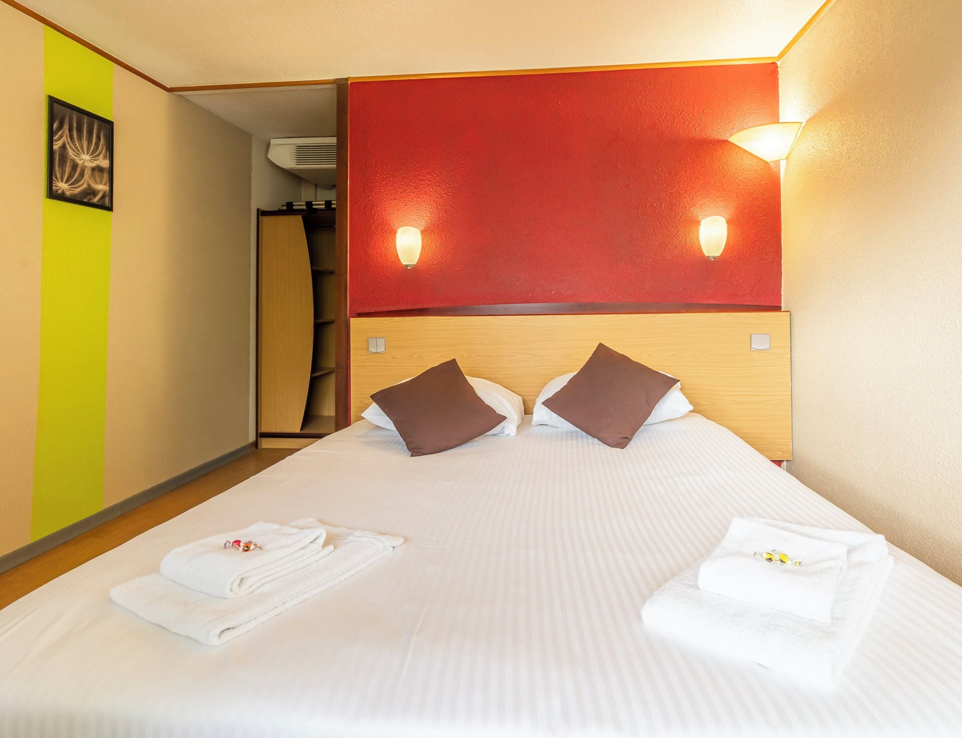 Habitación de hotel con cama, dos almohadas, mesita de noche y una pared roja decorativa.
