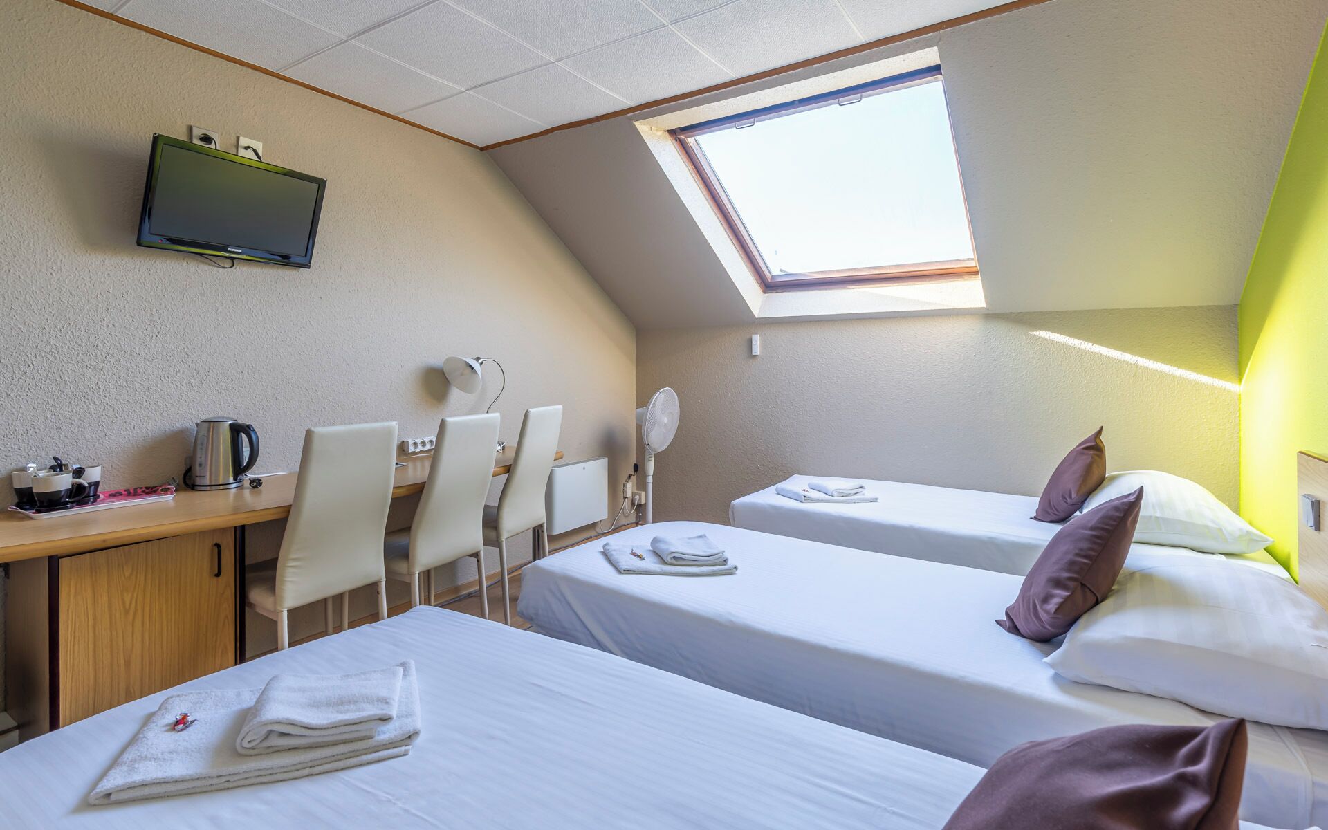 A hotel room with two beds, a desk with chairs, a television, and a skylight.