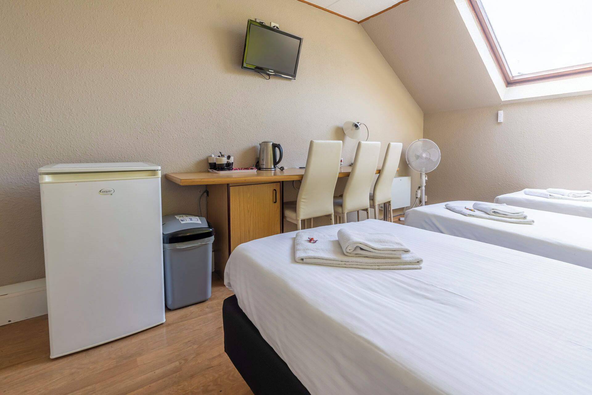 A hotel room with a desk, two chairs, a TV, a bed, and a small trash bin.