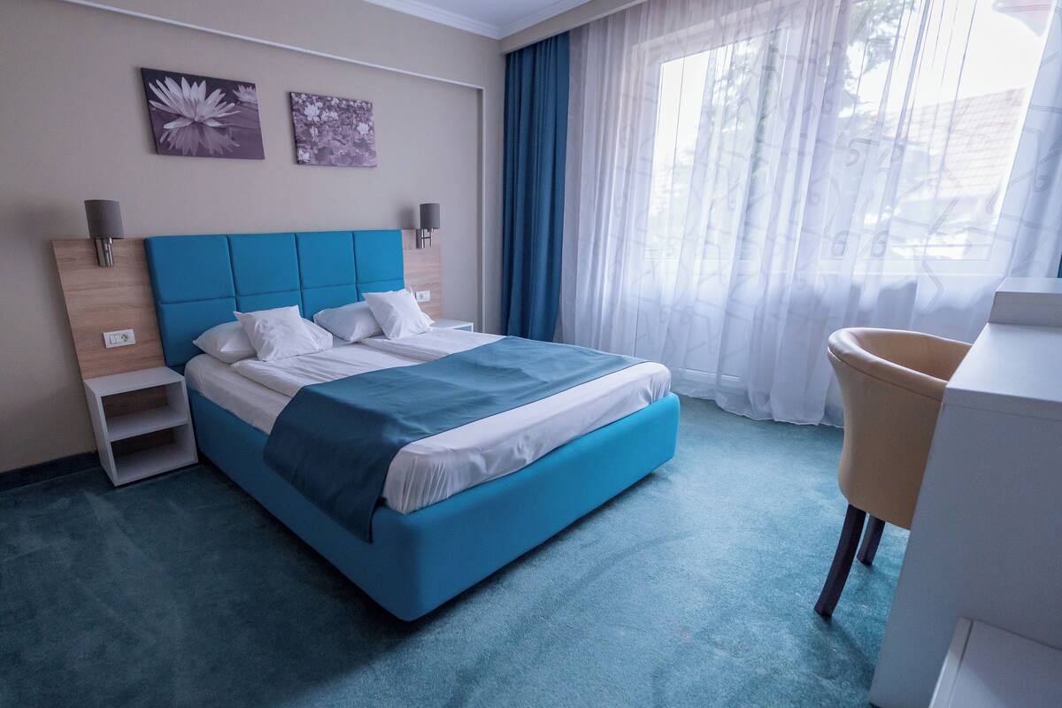 Egy modern szállodai szoba, amelyben egy nagy ágy, egy éjjeliszemély, egy szék és egy ablak található, melyen könnyű, áttetsző függöny van.