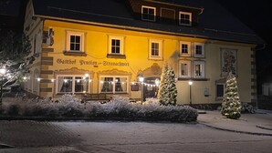 Un edificio con un letrero que dice "Gasthof Pension Strachwitz" de noche, mostrando un suelo cubierto de nieve y dos árboles de Navidad decorados.