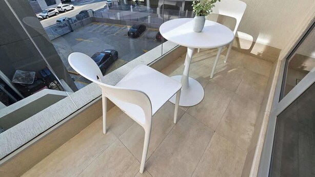 Un balcon avec une table et des chaises blanches, un pot de fleurs et une vue sur des voitures garées et un immeuble.