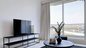 A modern living room with a flat-screen TV on a black TV stand, a round coffee table with a vase, and a large window with a view of a cityscape. - Luxury Apt W Pool Near Airport & Downtown Dubai (Dubai)