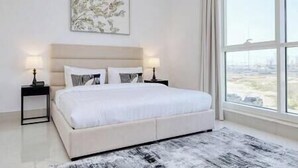 A bedroom with a large bed, two bedside tables with lamps, a window with a view, and a painting on the wall. - Luxury Apt W Pool Near Airport & Downtown Dubai (Dubai)